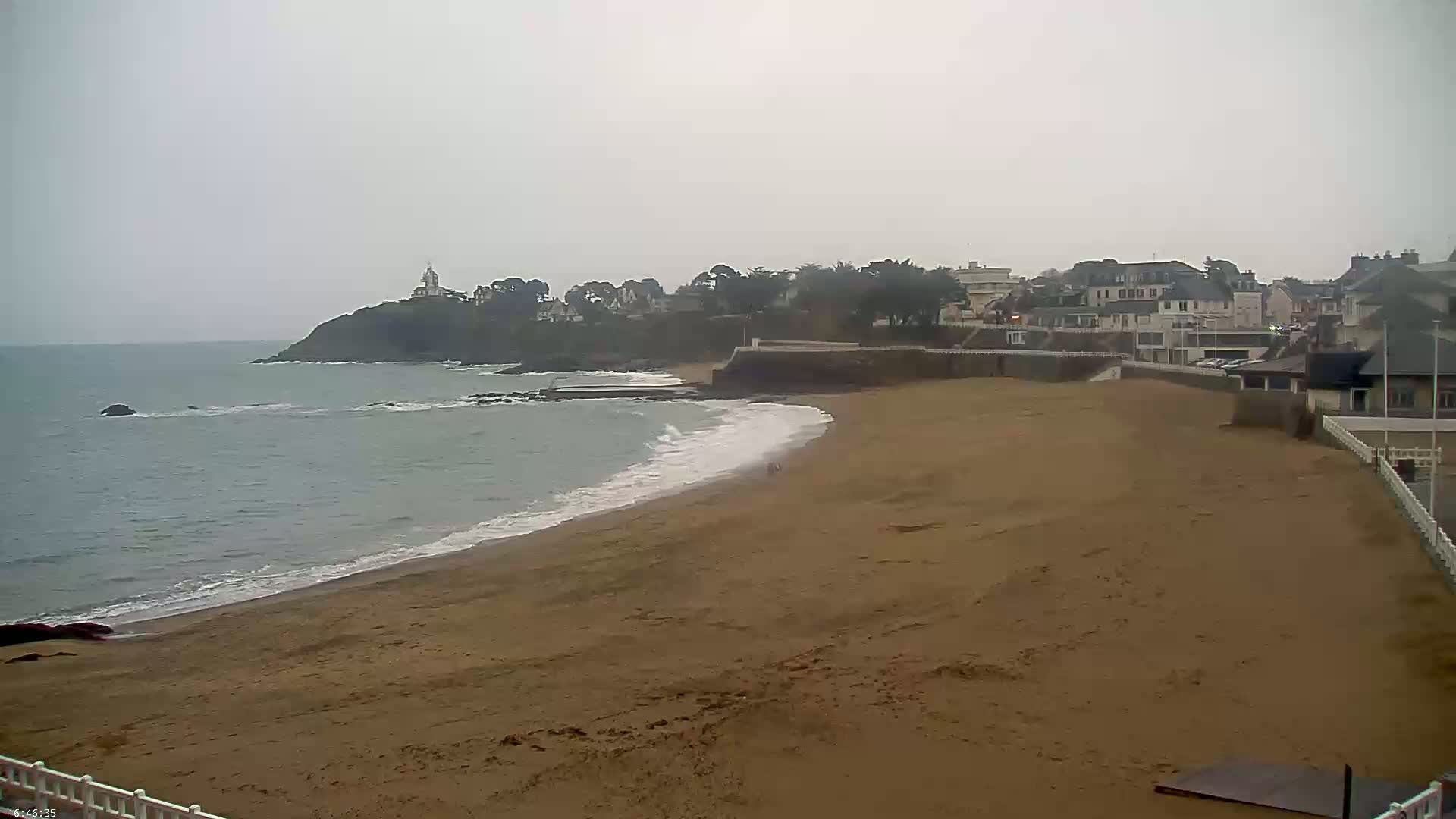 A sandy beach stretches along a calm, grey ocean under an overcast sky, with buildings and a lighthouse visible on a cliff in the background.