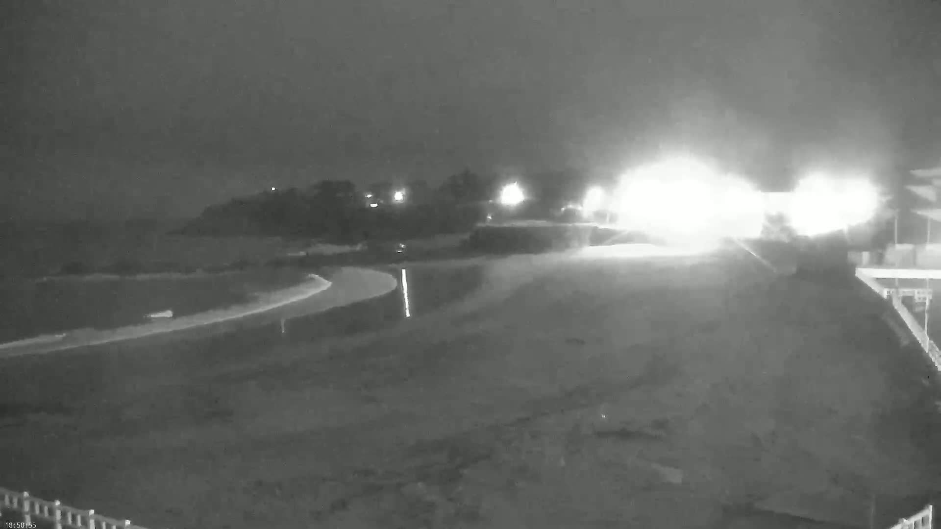 A nighttime, low-light view of a dark, sandy beach curving to the left, with dimly lit land and bright lights in the distance.