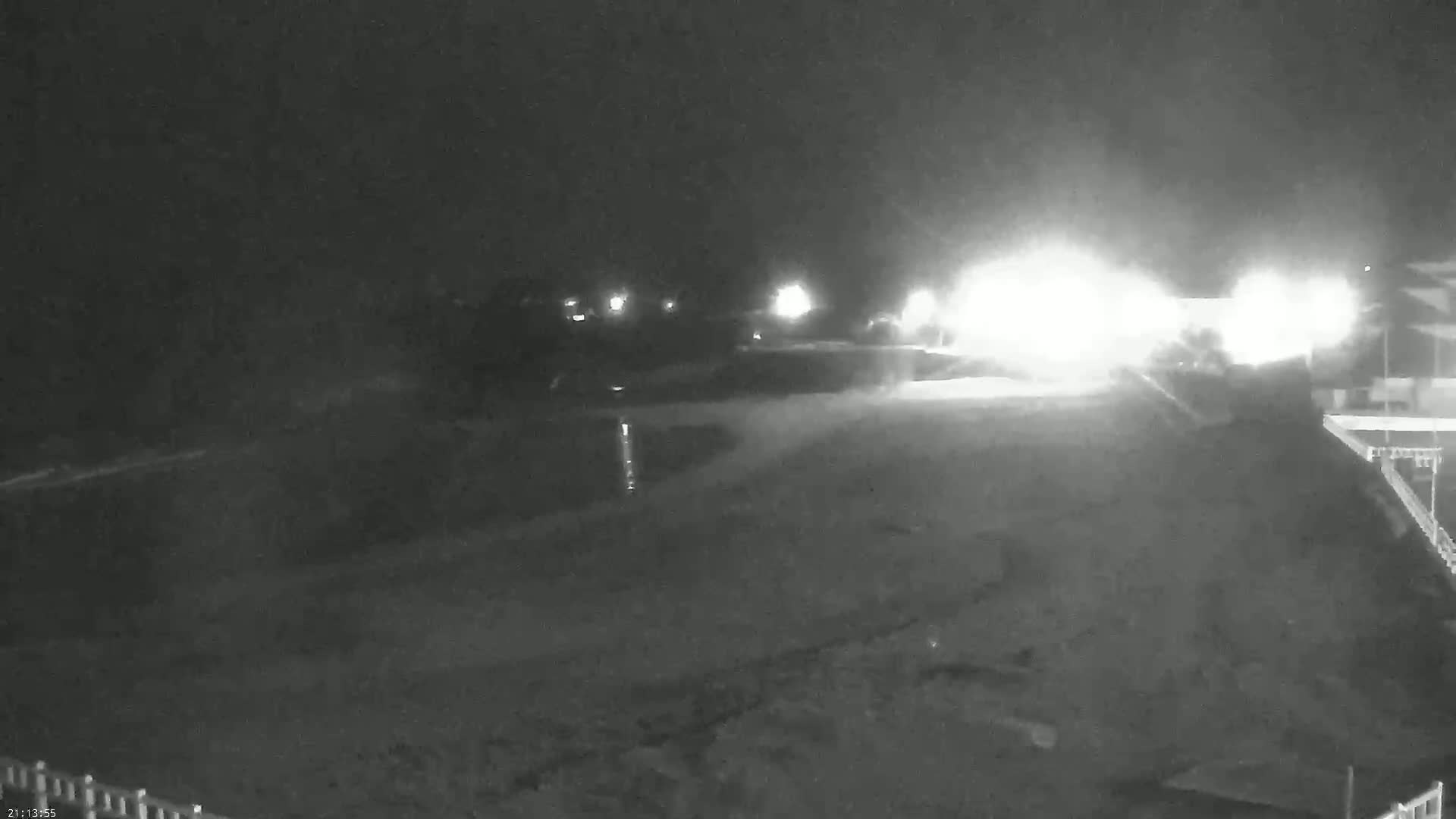 A nighttime, low-light image shows a dark, flat expanse of land with bright lights in the distance and a portion of a structure on the lower right.