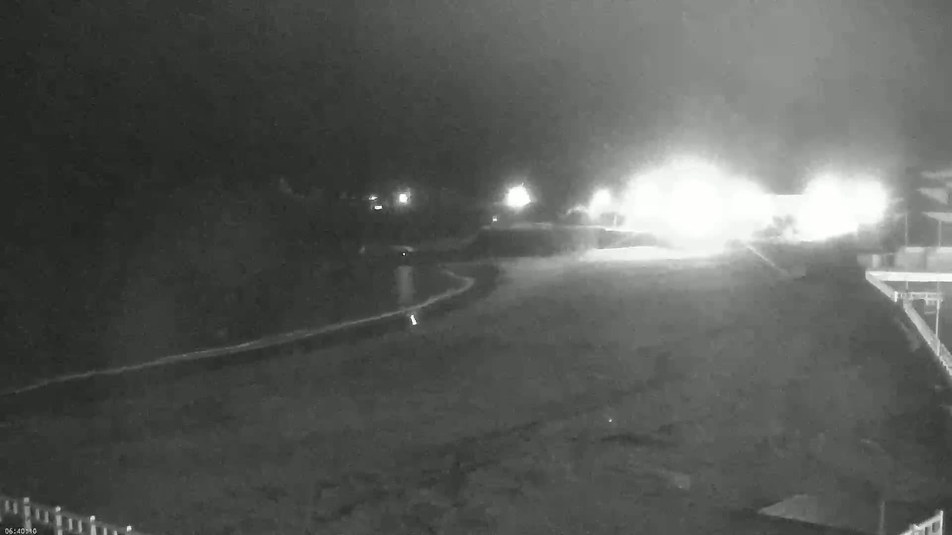 A nighttime, low-light view shows a dark, empty area with bright lights in the distance, possibly from vehicles, under a hazy or foggy sky.
