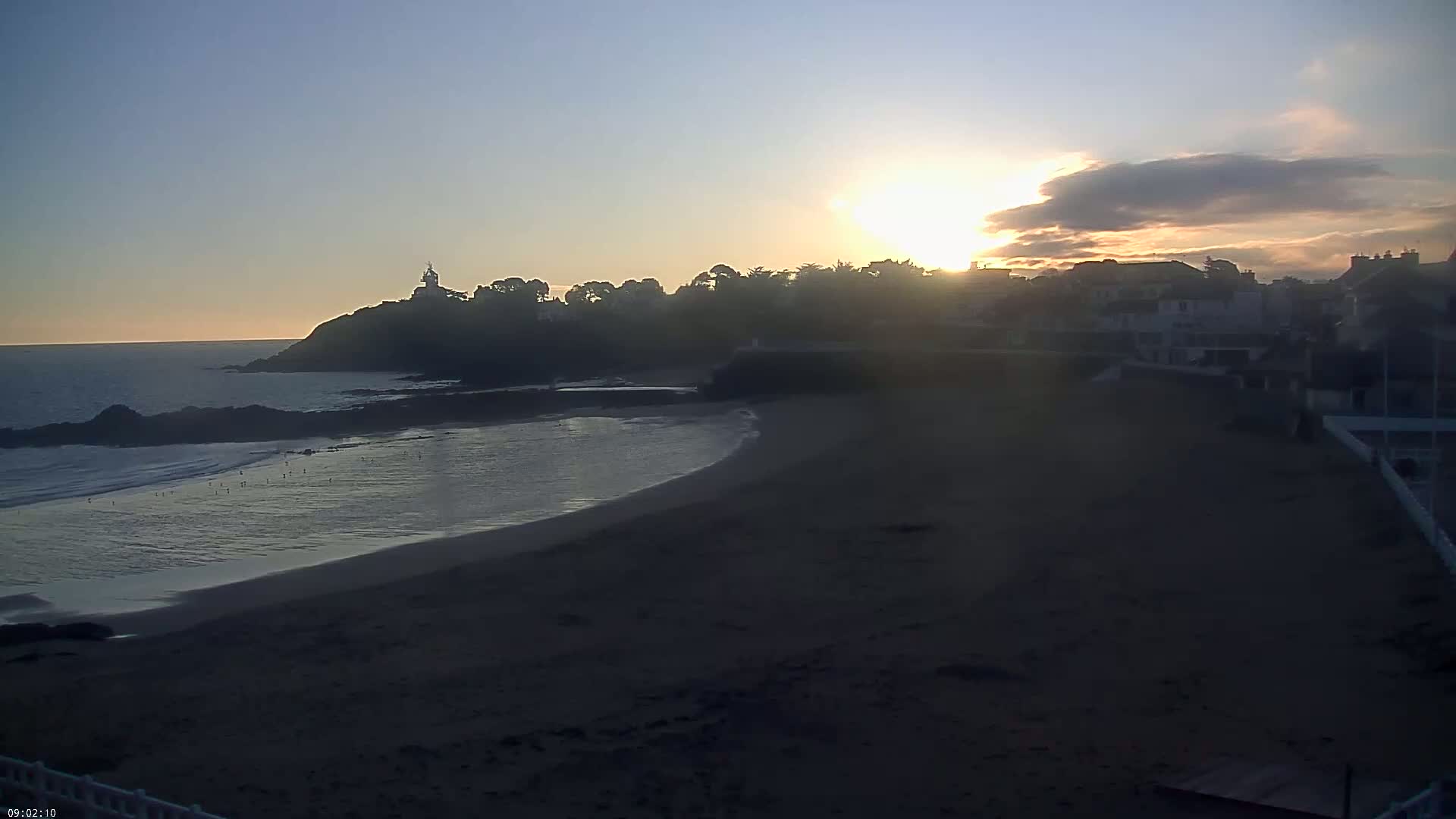 A sandy beach curves around a rocky point, with a town and a lighthouse silhouetted against a hazy sunrise.