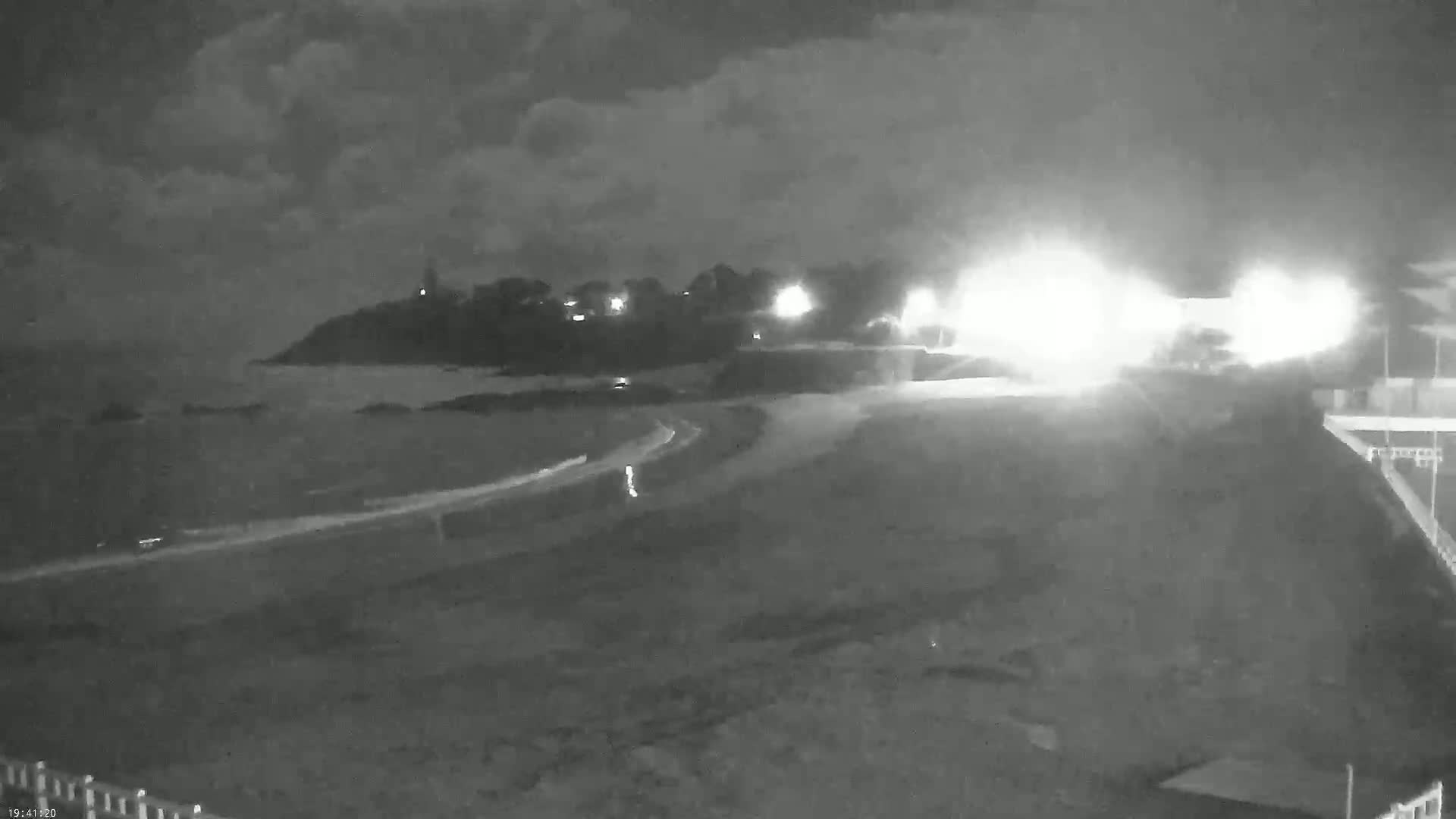 A nighttime, overcast view of a dark beach and coastline with lights visible on land and a small lighthouse in the distance.