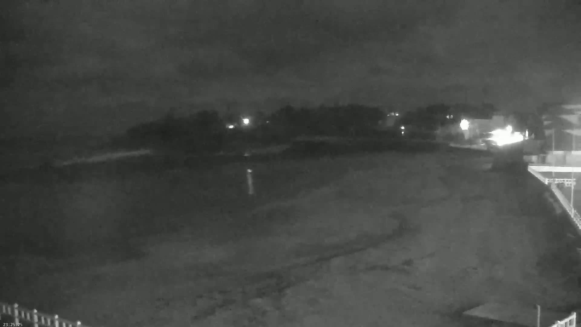 A nighttime, low-light view shows a dark beach or shoreline with a dimly lit area of trees and buildings in the background under a cloudy sky.