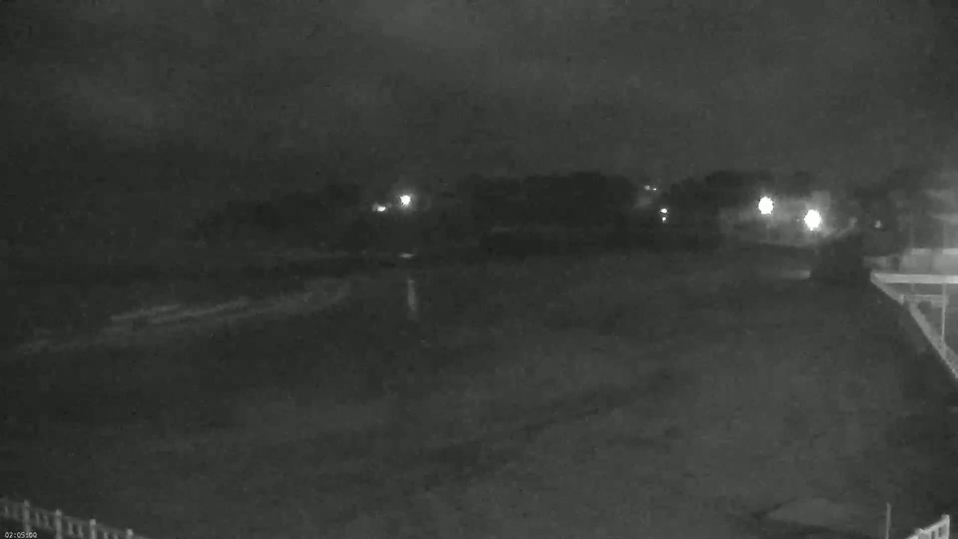 A nighttime, low-light view of a dark, flat expanse of land with a line of trees and distant lights in the background under an overcast sky.