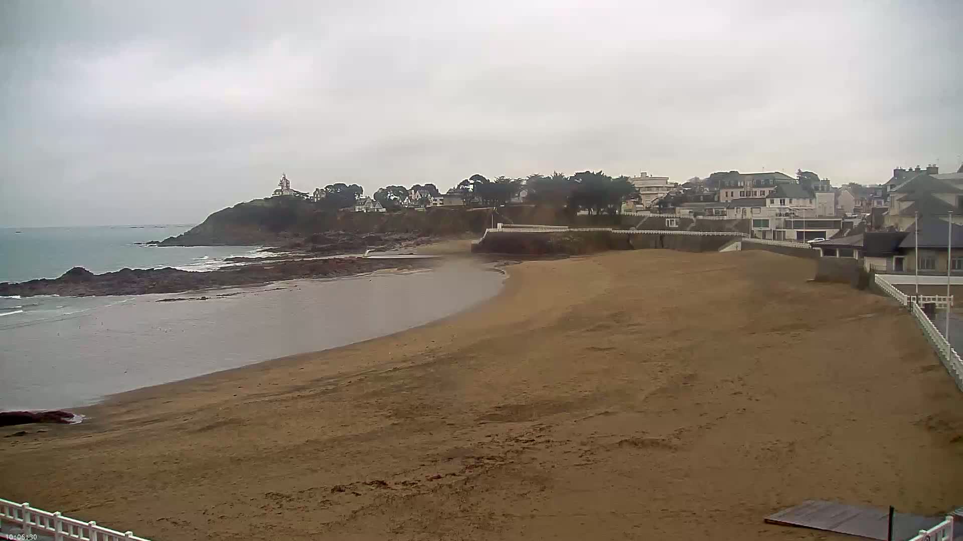 A sandy beach curves around a calm bay, with a small town and a lighthouse visible on a hill in the background under an overcast sky.