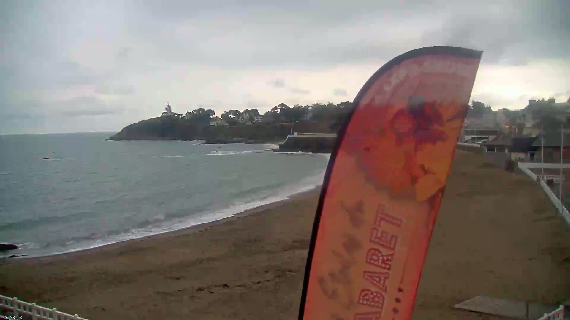A sandy beach stretches along a calm sea under an overcast sky, with a distant headland featuring buildings and trees.