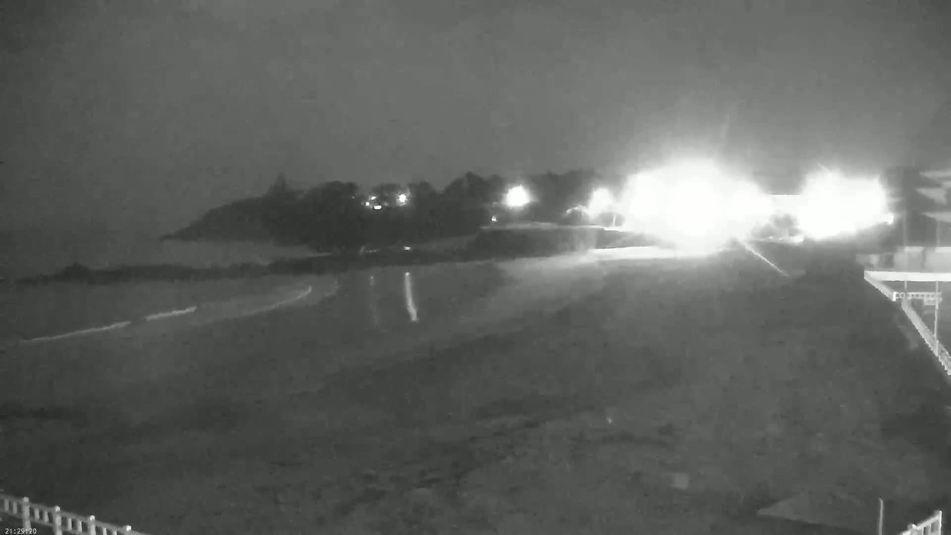 A nighttime, low-light view shows a dark beach and shoreline with some lights visible in the distance, and what appears to be wet pavement in the foreground.