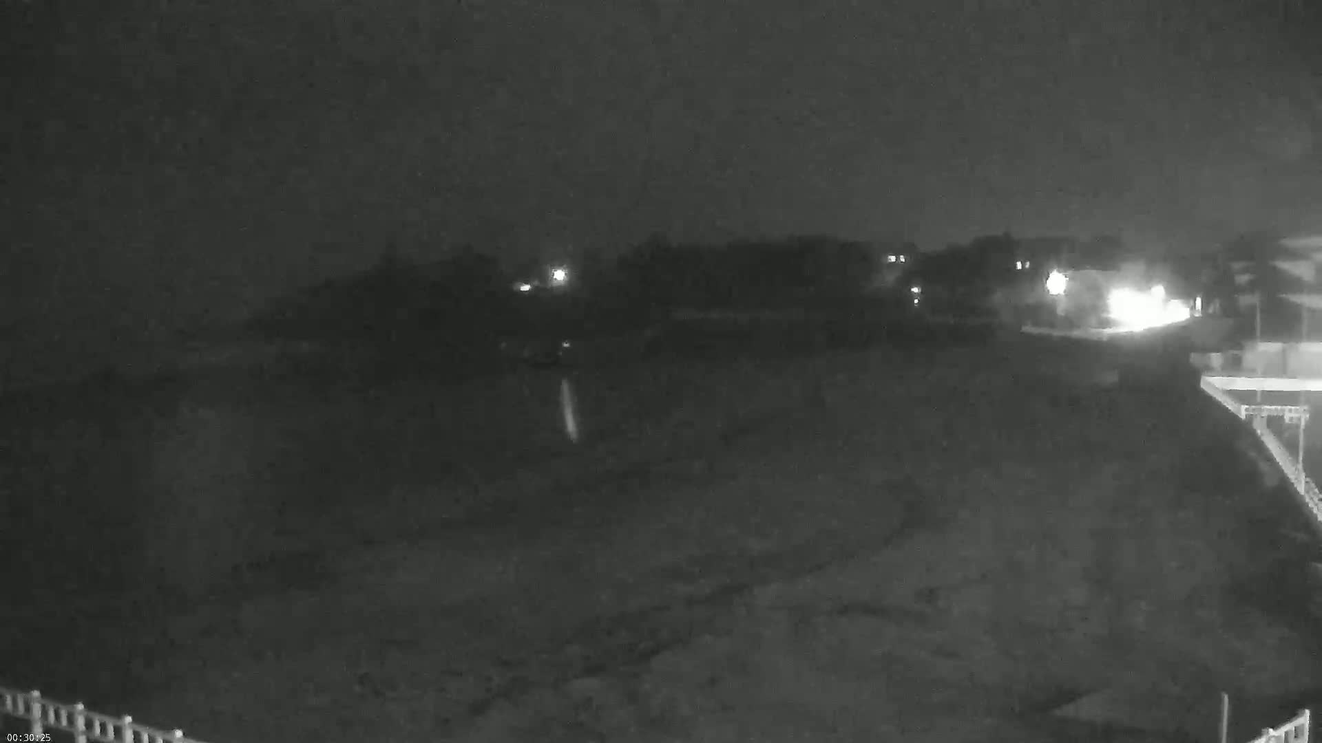 A nighttime, low-light view shows a dark, mostly empty expanse of land with some distant lights, and a section of a building or structure at the bottom right.
