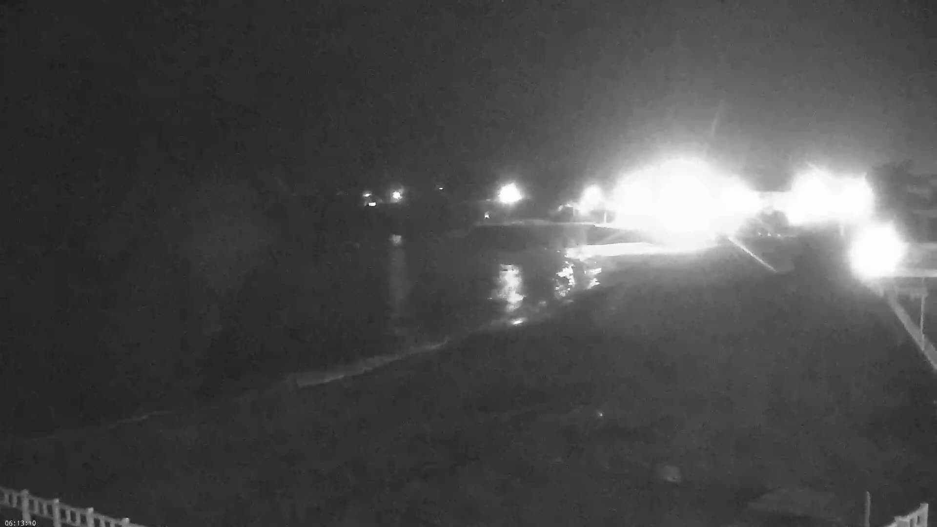 A nighttime, low-light view shows a dark body of water reflecting bright lights from a roadway beyond.