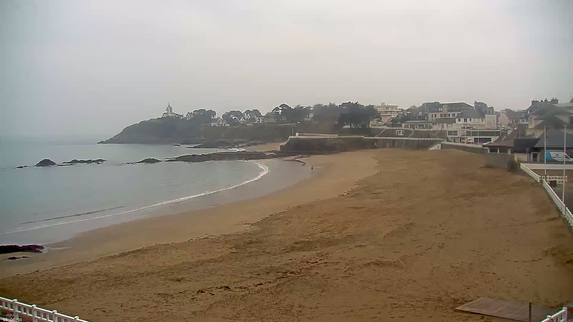 A sandy beach curves along a calm ocean under an overcast sky, with a small town and a distant lighthouse visible on a cliff in the background.