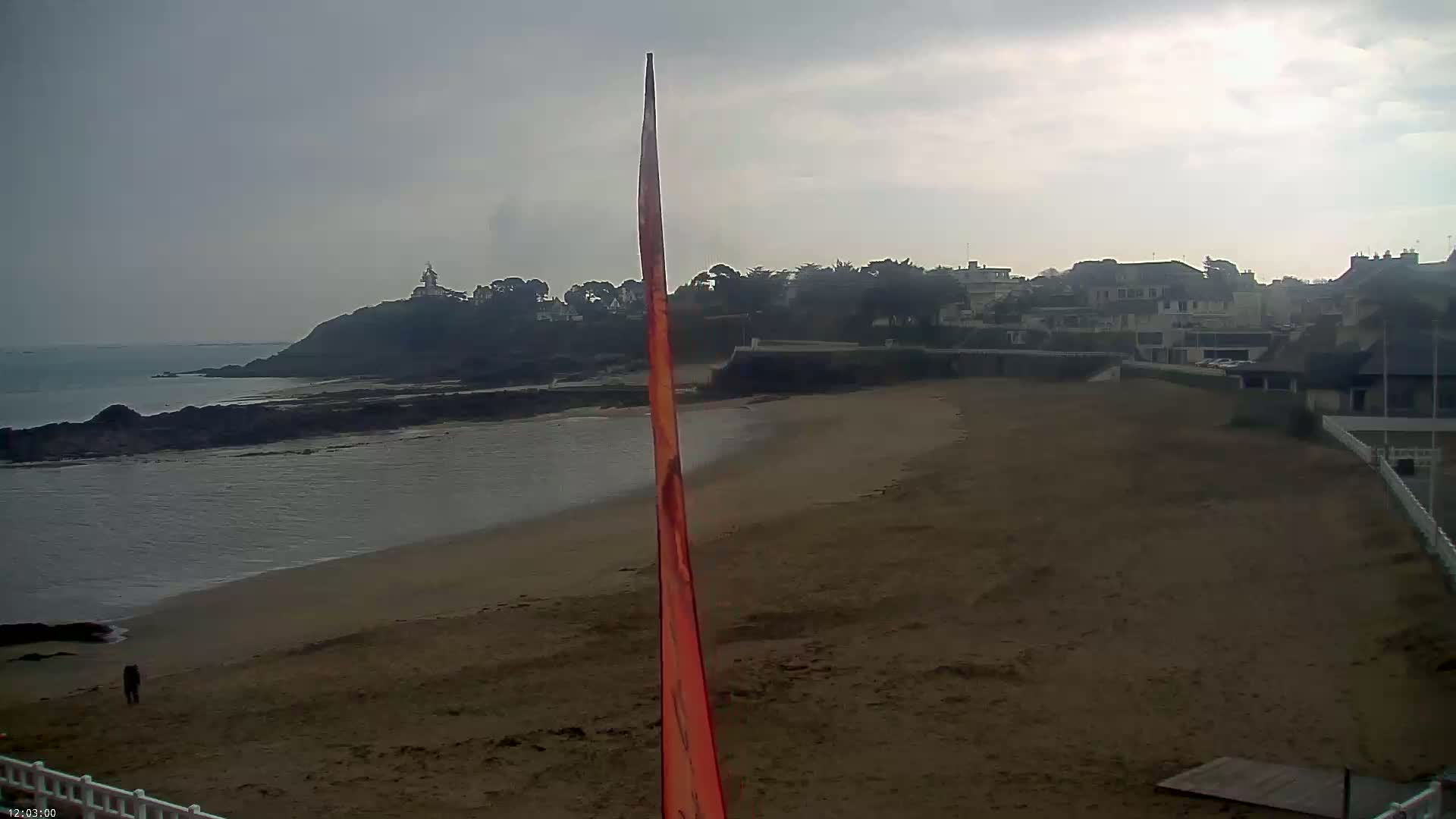 A sandy beach stretches out to a calm sea under an overcast sky, with a small town and a lighthouse visible in the distance.