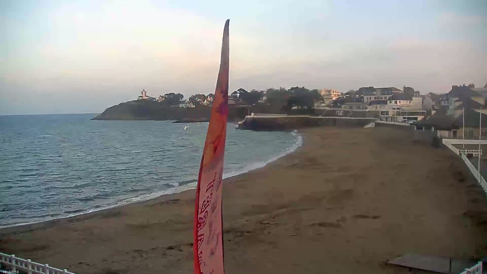 A sandy beach stretches out to a calm sea under a pale sky, with a small town and a lighthouse visible in the distance.