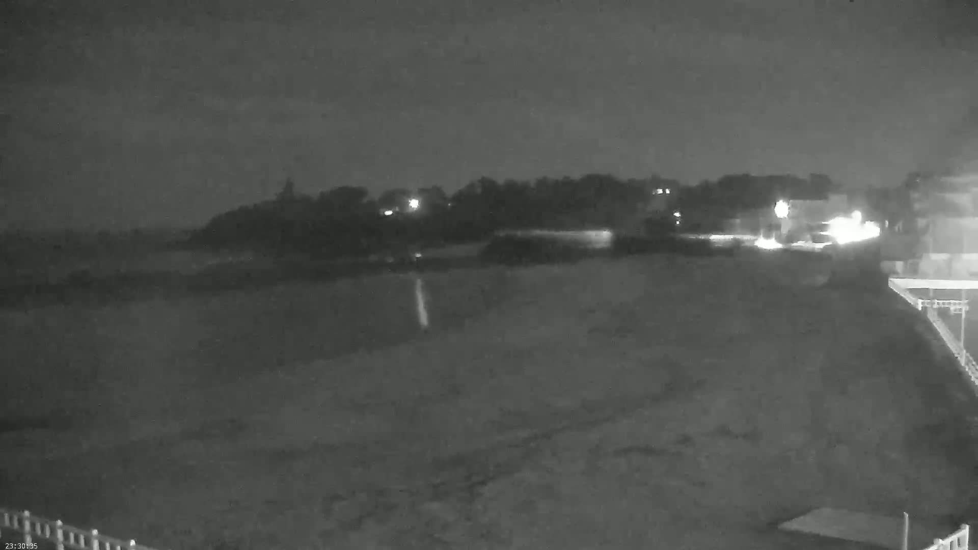 A nighttime, low-light view shows a dark, flat expanse of land leading to a treeline with a few distant lights under a cloudy sky.