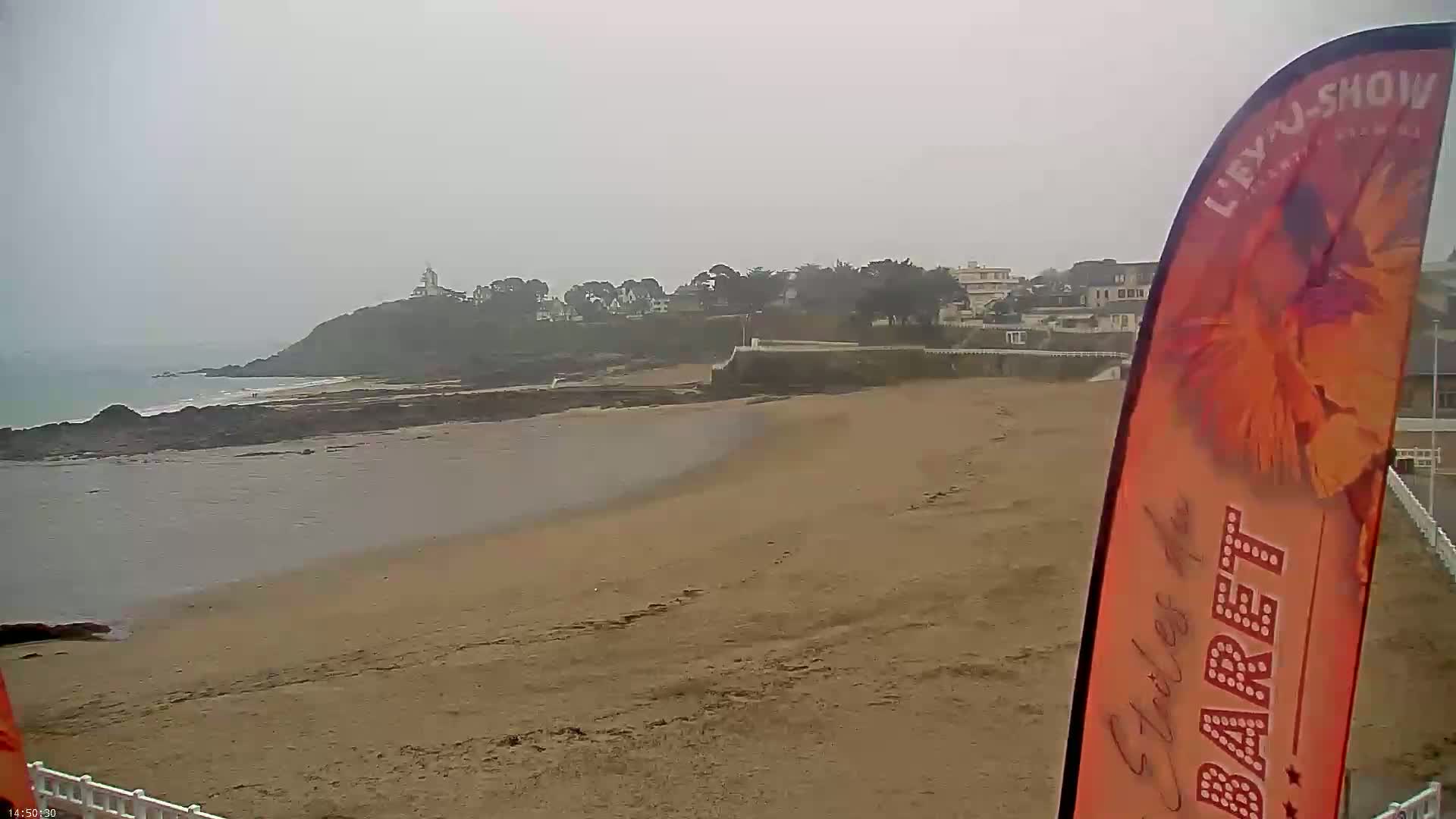 A sandy beach stretches out to a calm ocean under an overcast sky, with a small town and a lighthouse visible on a cliff in the distance.