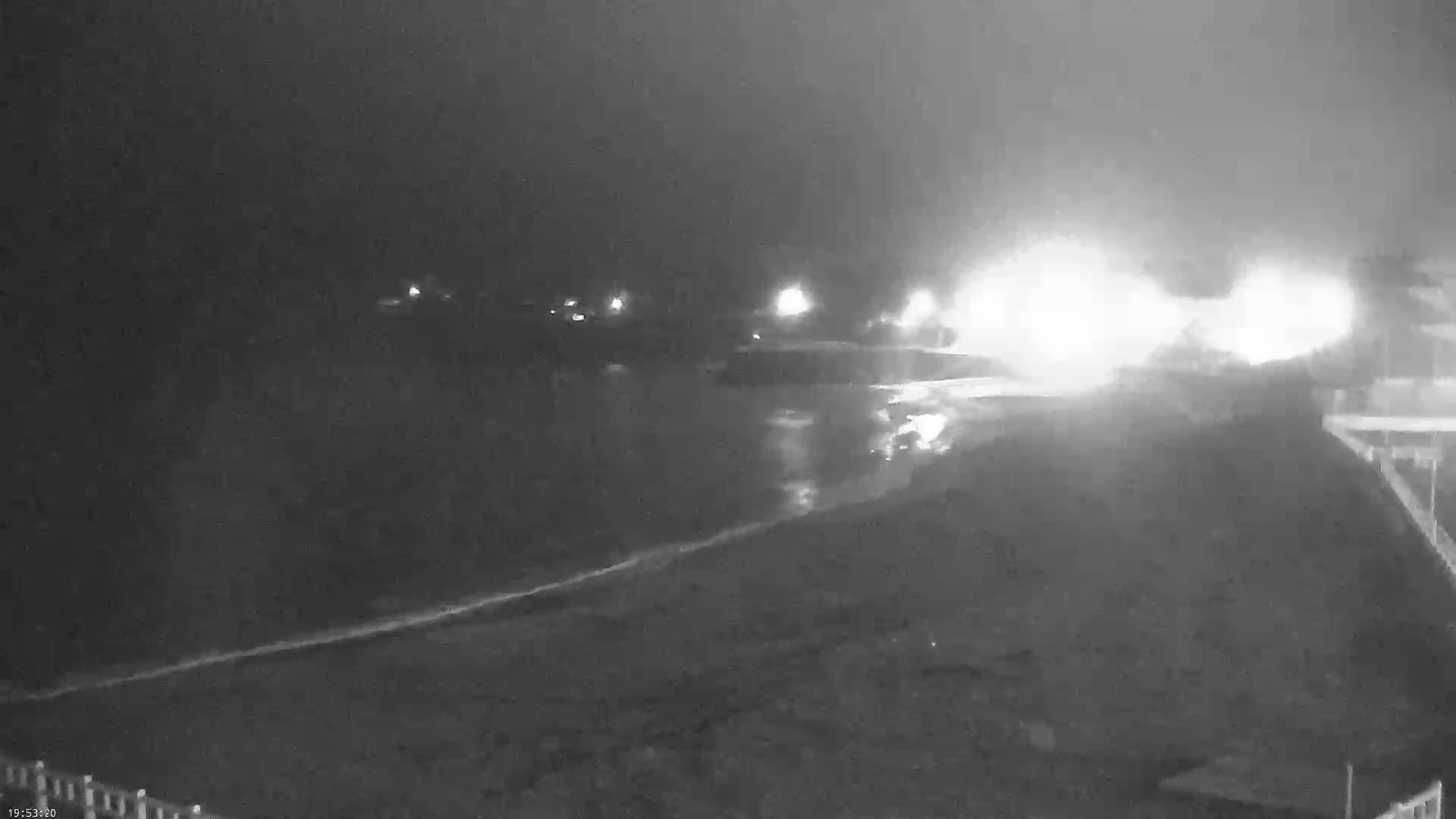 A nighttime, blurry, low-light view of a dark beach with a wavy shoreline, distant bright lights, and some structures visible on the right.