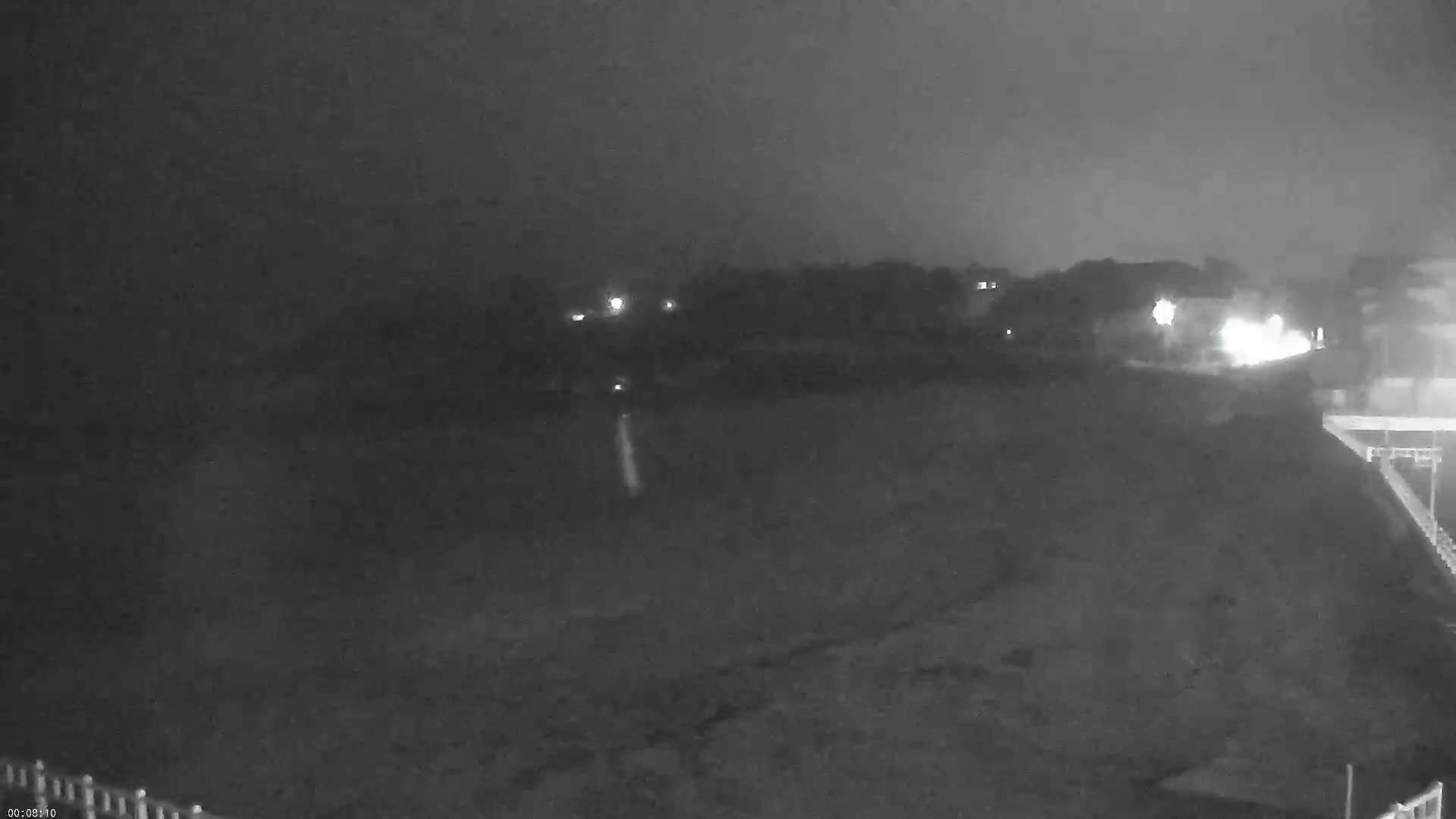 A nighttime, low-light view shows a dark, flat expanse possibly a beach or field, with a dimly lit shoreline and distant lights in the background under an overcast sky.