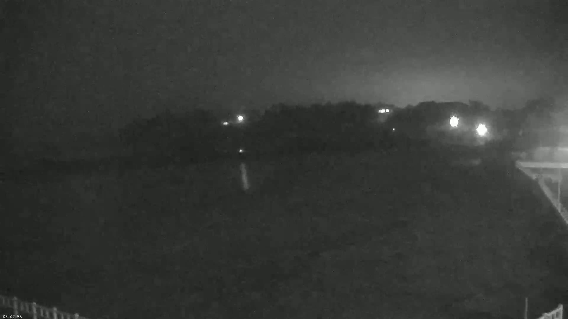 A nighttime, low-light view shows a dark, flat expanse of land with a line of trees and distant lights in the background.