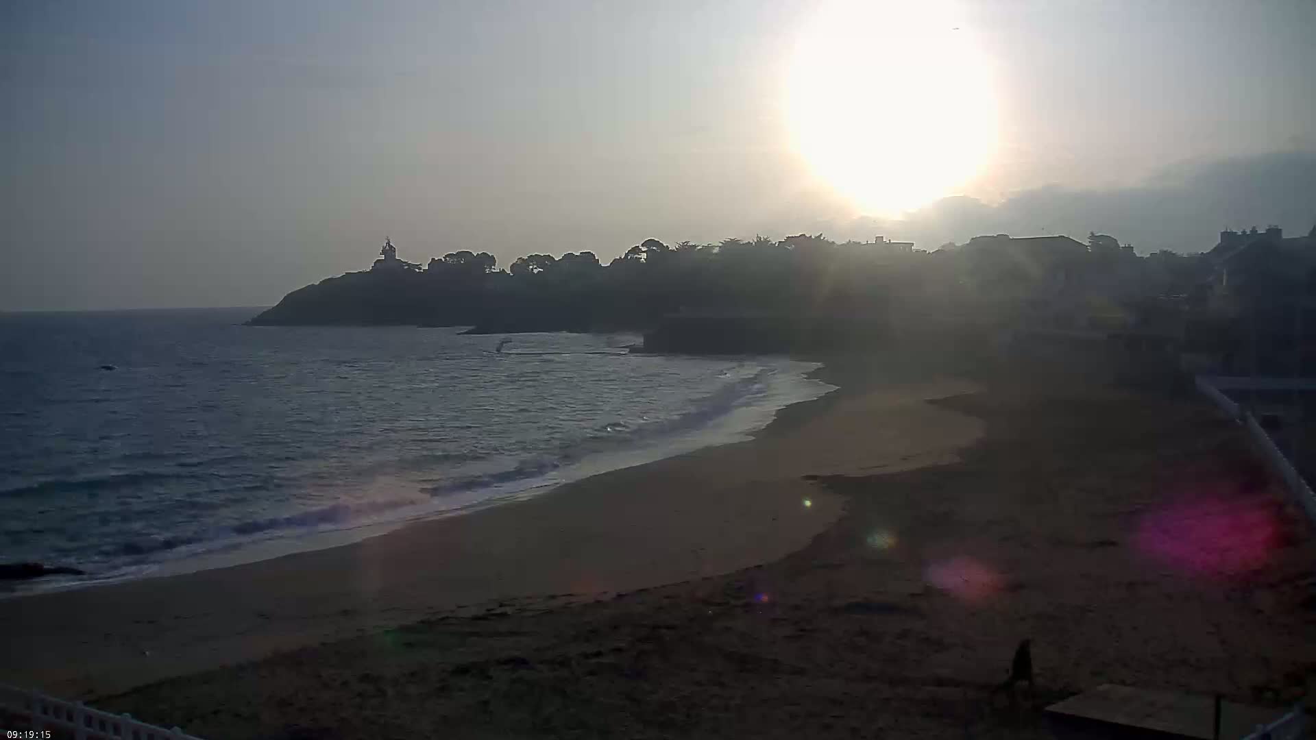 A sandy beach curves gently toward a hazy sunset over calm ocean waves, backed by a tree-lined coastline with buildings and a small tower.
