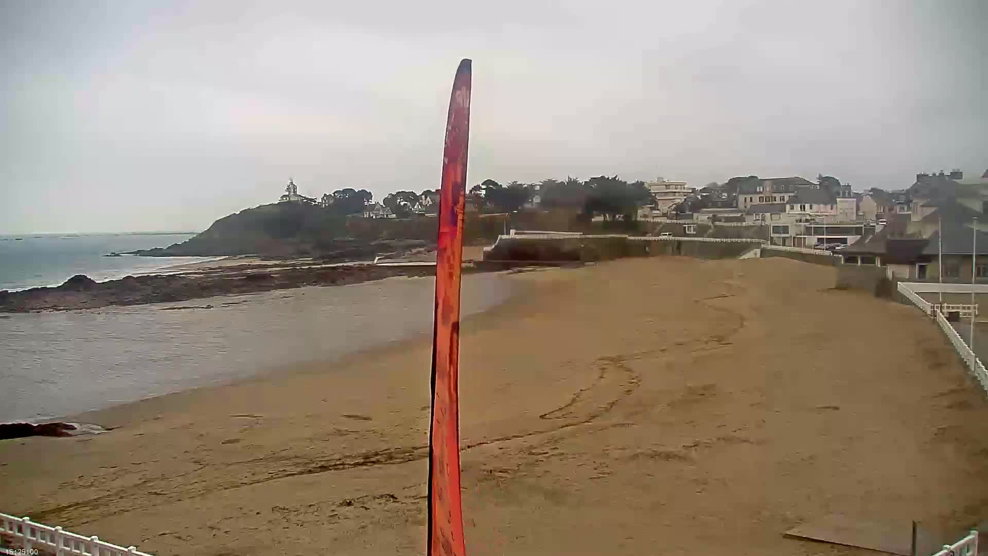 A long, thin, orange-red vertical object stands on a sandy beach overlooking a calm sea under an overcast sky, with buildings and a small hill visible in the distance.