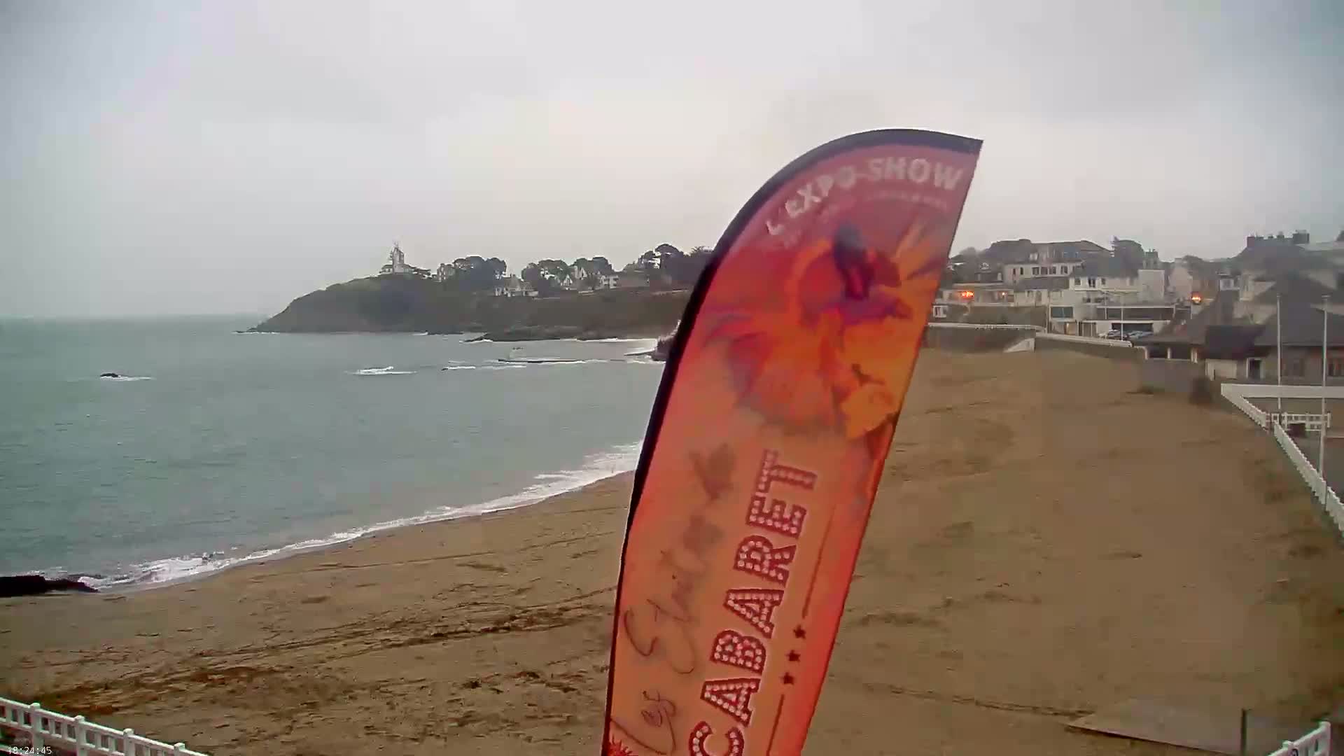 A sandy beach stretches toward a calm sea under an overcast sky, with a distant headland featuring a lighthouse and buildings visible beyond.