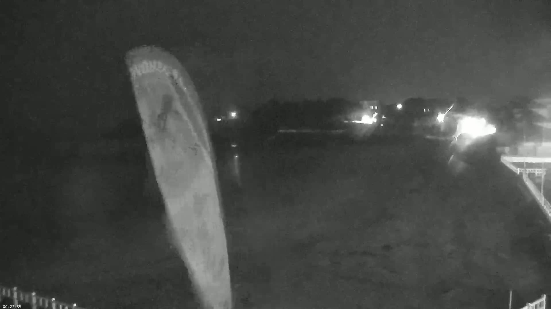 A nighttime, low-light grayscale image shows a large, light-colored object, possibly a sail or sheet, angled to the right, with a dark background showing indistinct lights in the distance.