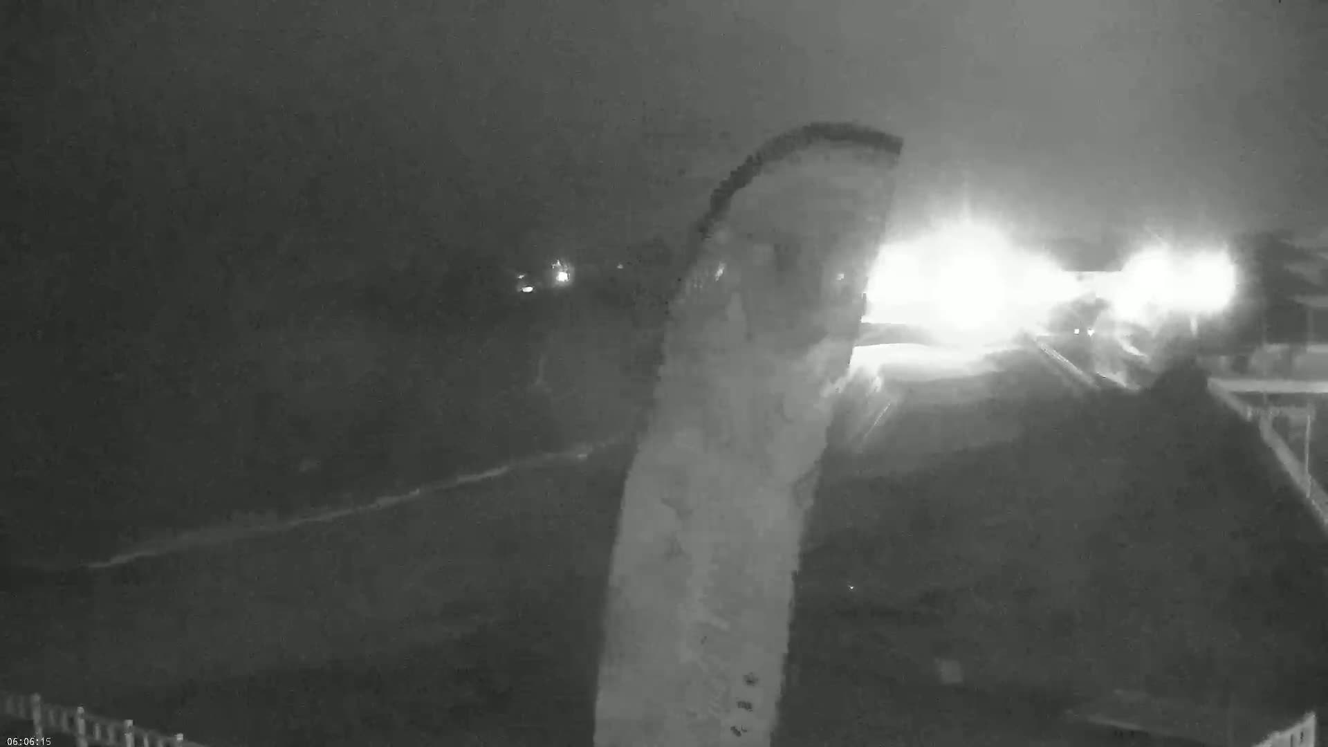 A nighttime, low-light view shows bright headlights from a vehicle approaching on a roadway, with other lights visible in the distance.