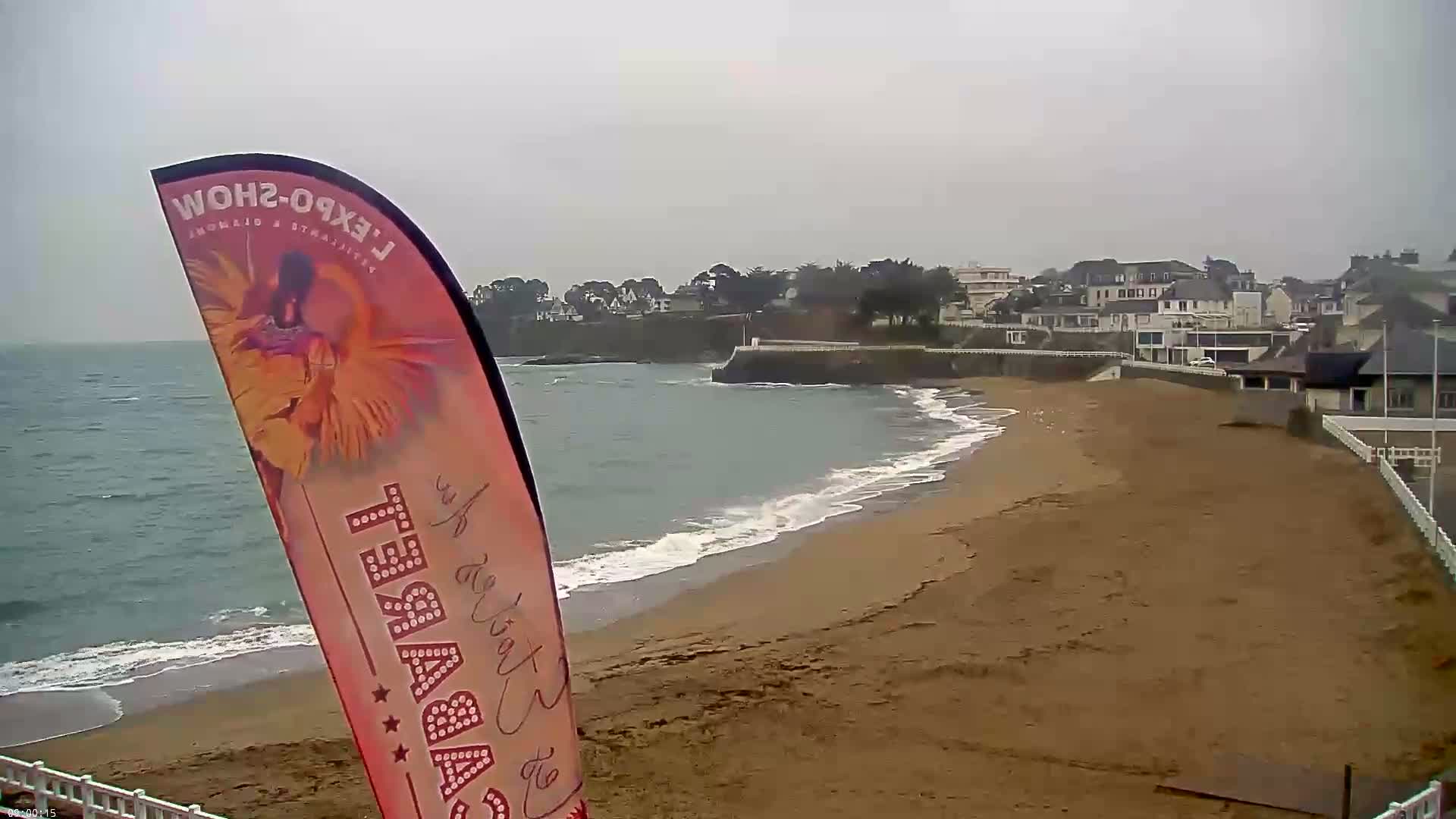 A sandy beach with gentle waves laps the shore under an overcast sky, next to a coastal town.