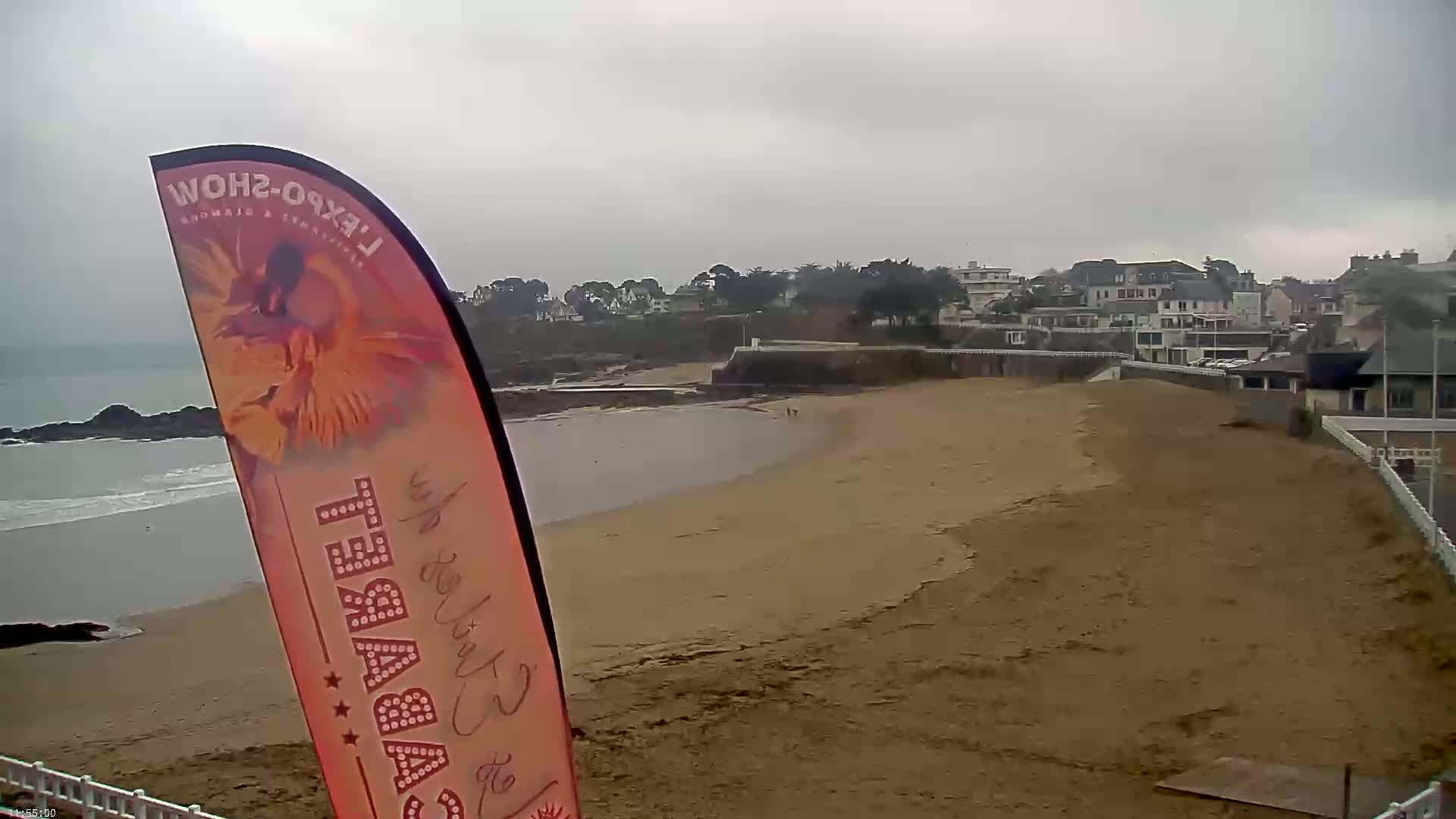 A sandy beach stretches out to a calm ocean under an overcast sky, with a small town visible in the background.