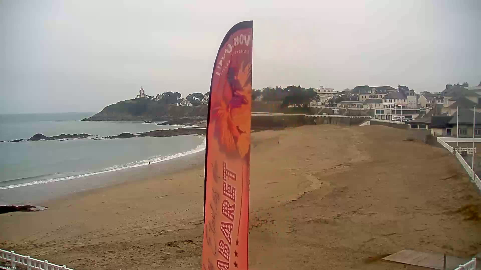A sandy beach stretches out to a calm sea under an overcast sky, with a small town and a rocky headland visible in the distance.