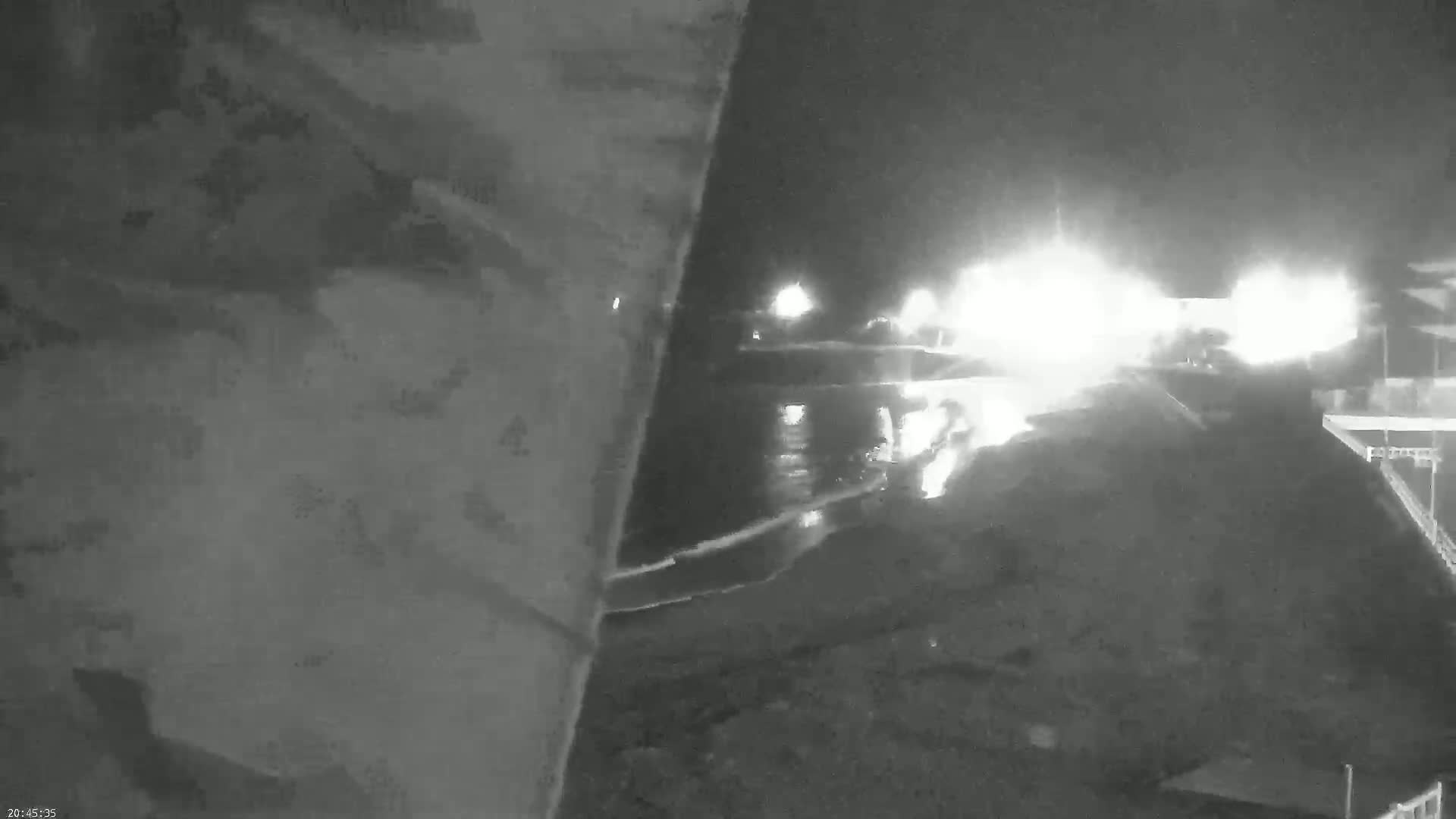 A nighttime, low-light view shows bright lights reflecting on a dark, wet surface near a structure, possibly a dock or pier.