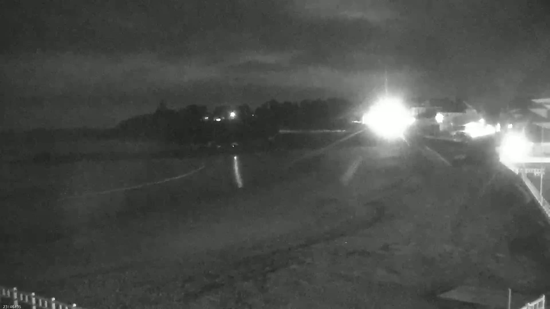 A nighttime, low-light view shows a dark beach or shoreline with faint lights from buildings and vehicles in the distance under a cloudy sky.