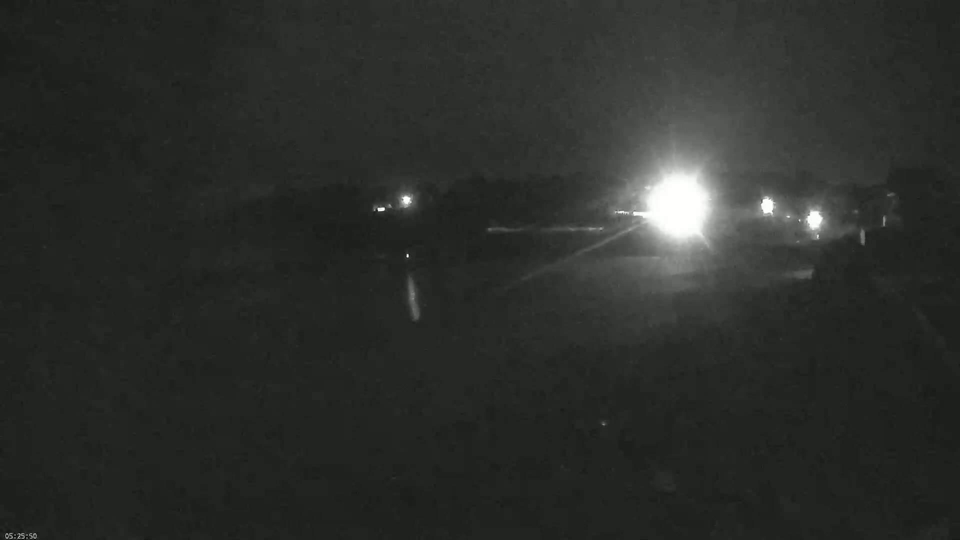 At night, vehicle headlights illuminate a dark road with dimly lit trees and buildings in the background.