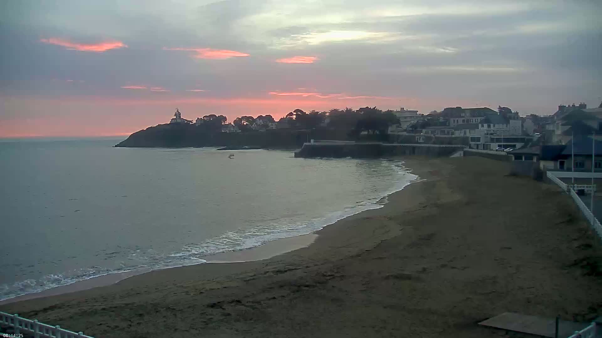 A sandy beach with gentle waves laps at the shore under a pinkish sunset sky, with houses and a small lighthouse perched on a nearby cliff.