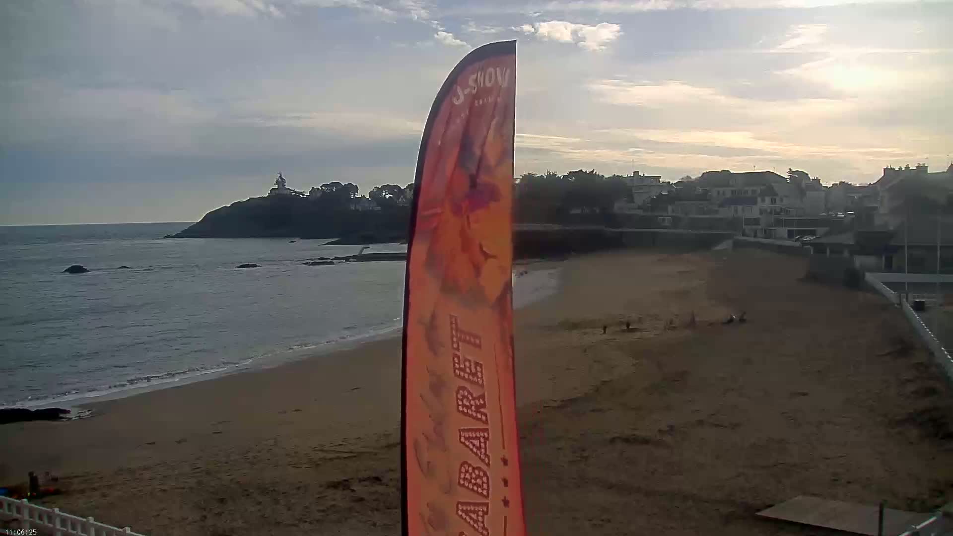 A sandy beach stretches toward a calm ocean under a partly cloudy sky, with a small town and a distant lighthouse visible in the background, and a tall orange flag standing prominently in the foreground.