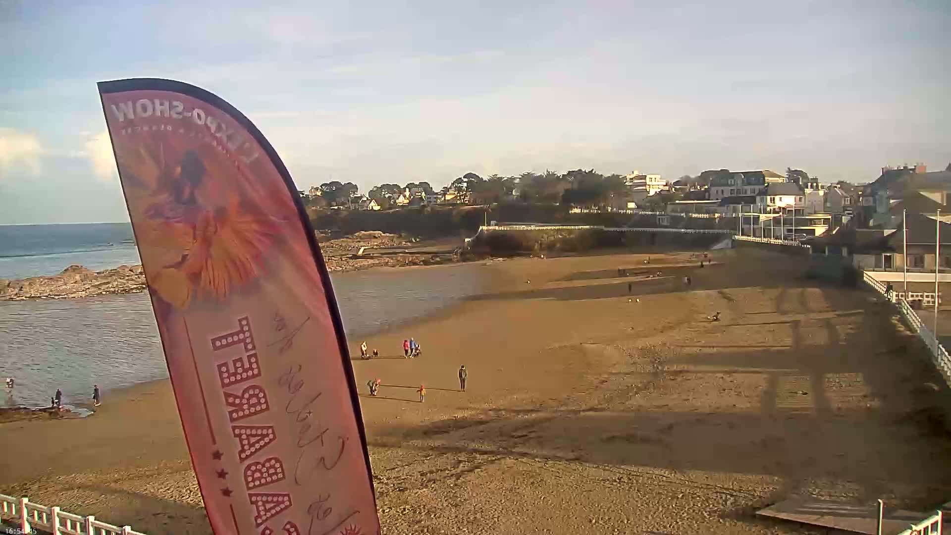 A sandy beach with a few people and buildings in the background, under a clear, bright sky.