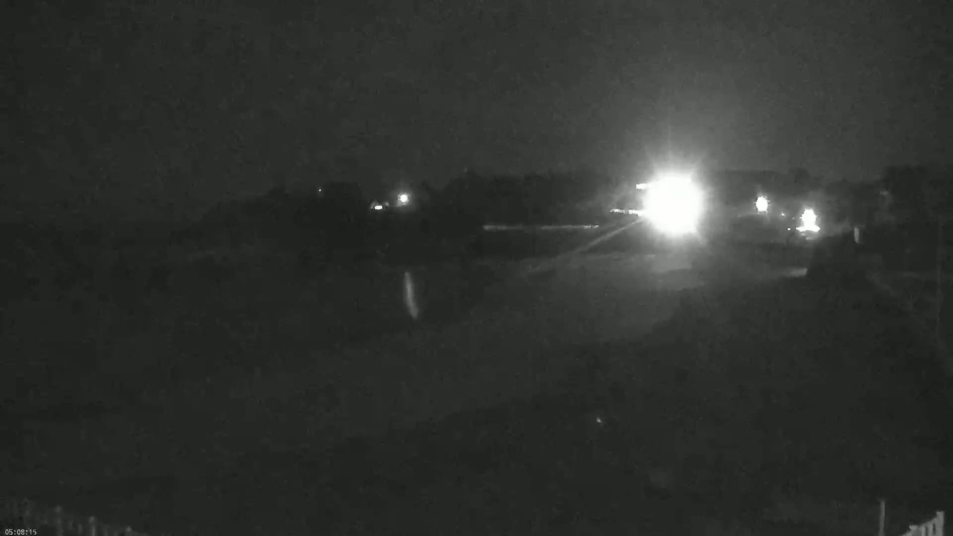 At night, bright headlights illuminate a dark road and the surrounding landscape, which appears to be mostly vegetation.