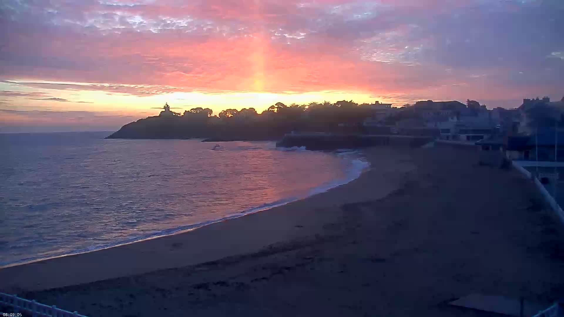 A sandy beach curves along a coastline under a colorful sunset sky, with calm waves lapping the shore and buildings silhouetted against the light.