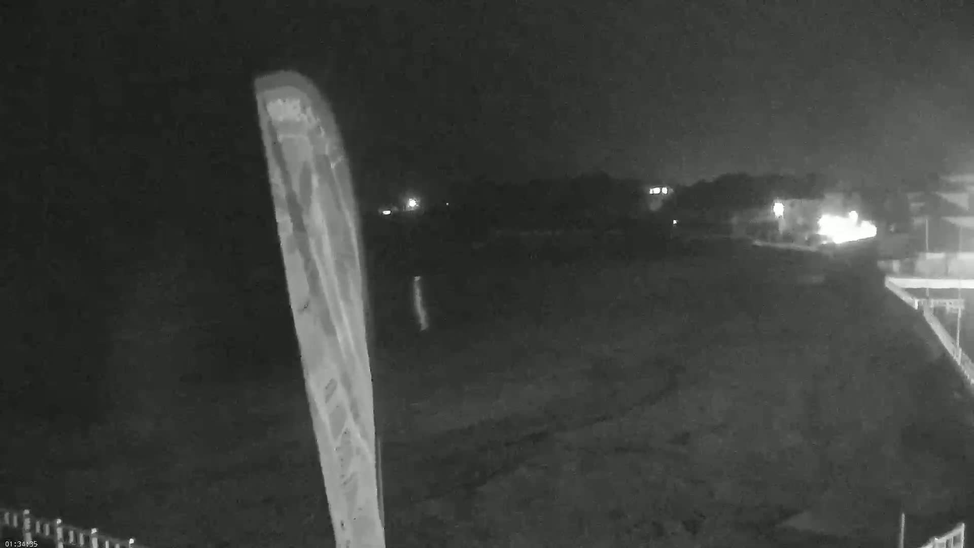 A nighttime, low-light image shows a dark, flat expanse of land with a tall, thin object in the left foreground, and a few distant lights visible in the background.