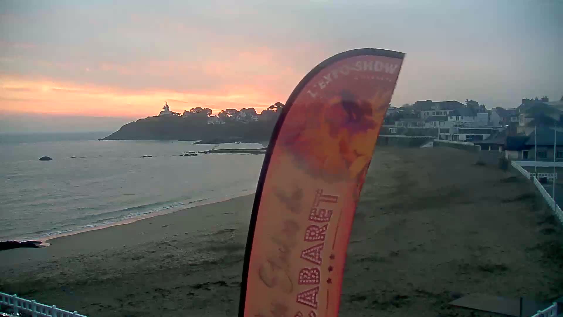 A sandy beach at sunset overlooks calm ocean waters and a distant town nestled on a point of land with a lighthouse.