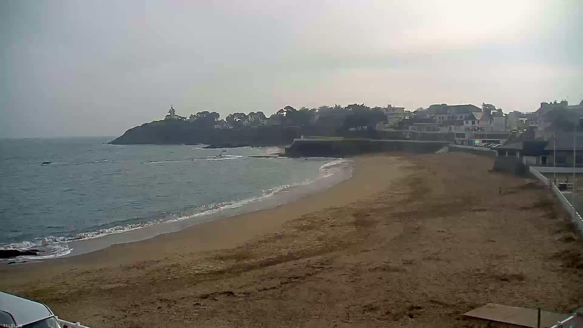 A sandy beach stretches along a calm, gray ocean under an overcast sky, with a town and a lighthouse visible in the distance on a rocky headland.