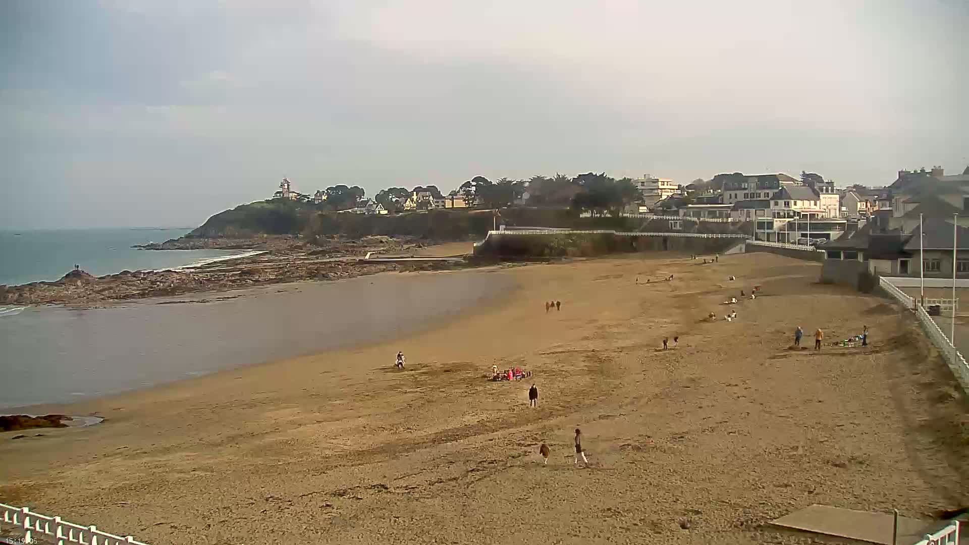 A sandy beach with scattered people extends from a calm body of water toward a town nestled on a cliff under an overcast sky.