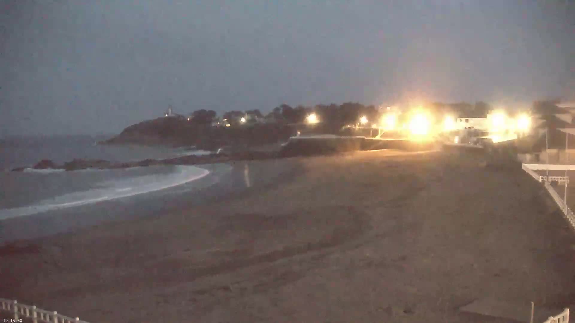 A dark, sandy beach at night is illuminated by lights from buildings and structures on the shore, with calm waves lapping at the sand and a distant headland visible under a dusky sky.