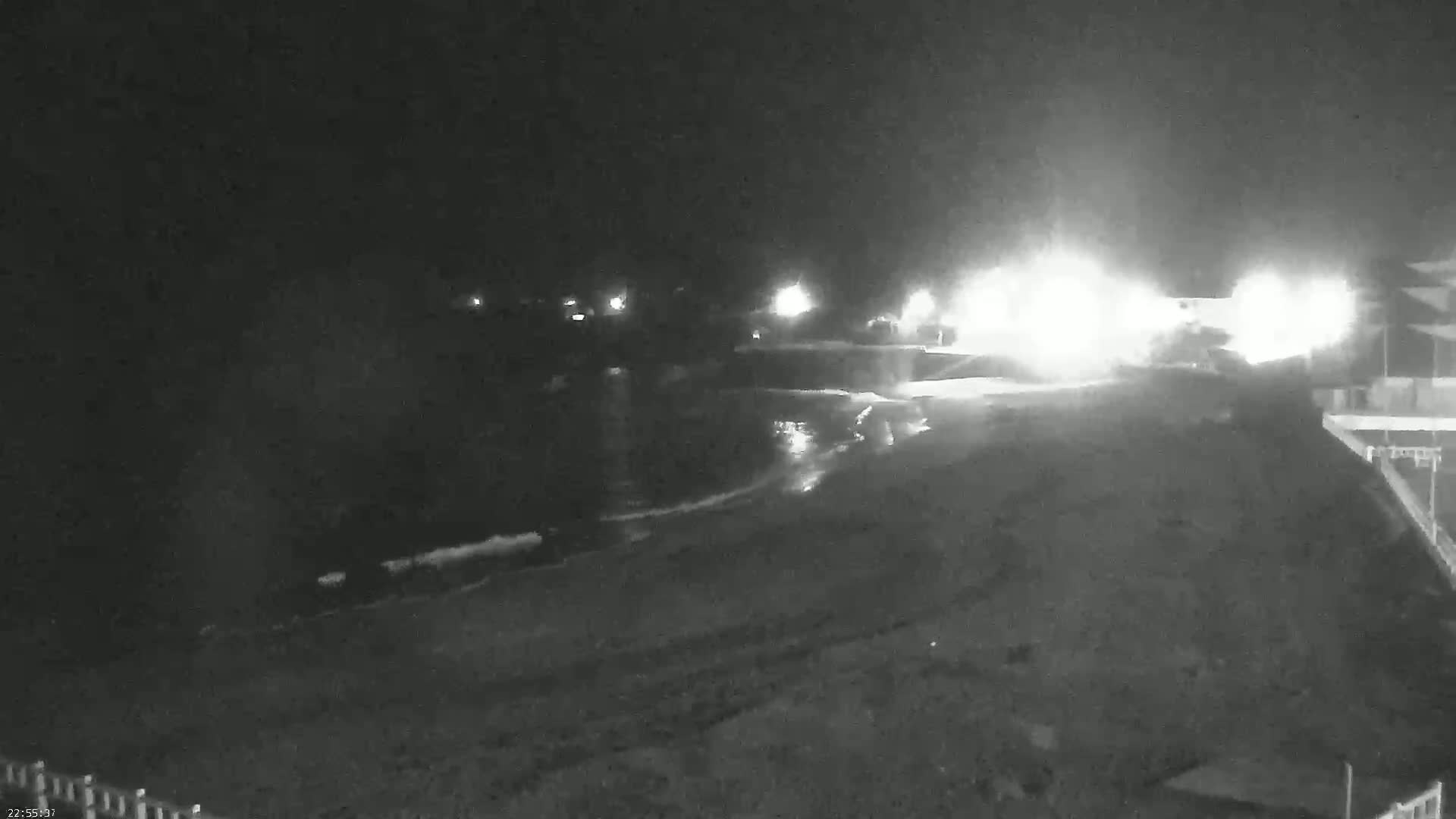 A nighttime, low-light view of a dark beach with waves lapping the shore, illuminated by bright lights in the distance.
