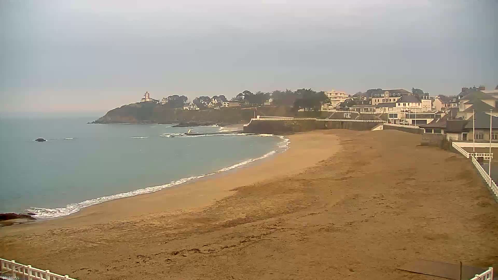 A sandy beach stretches along a calm, gray ocean under an overcast sky, bordered by a cliff with buildings and a lighthouse in the distance.