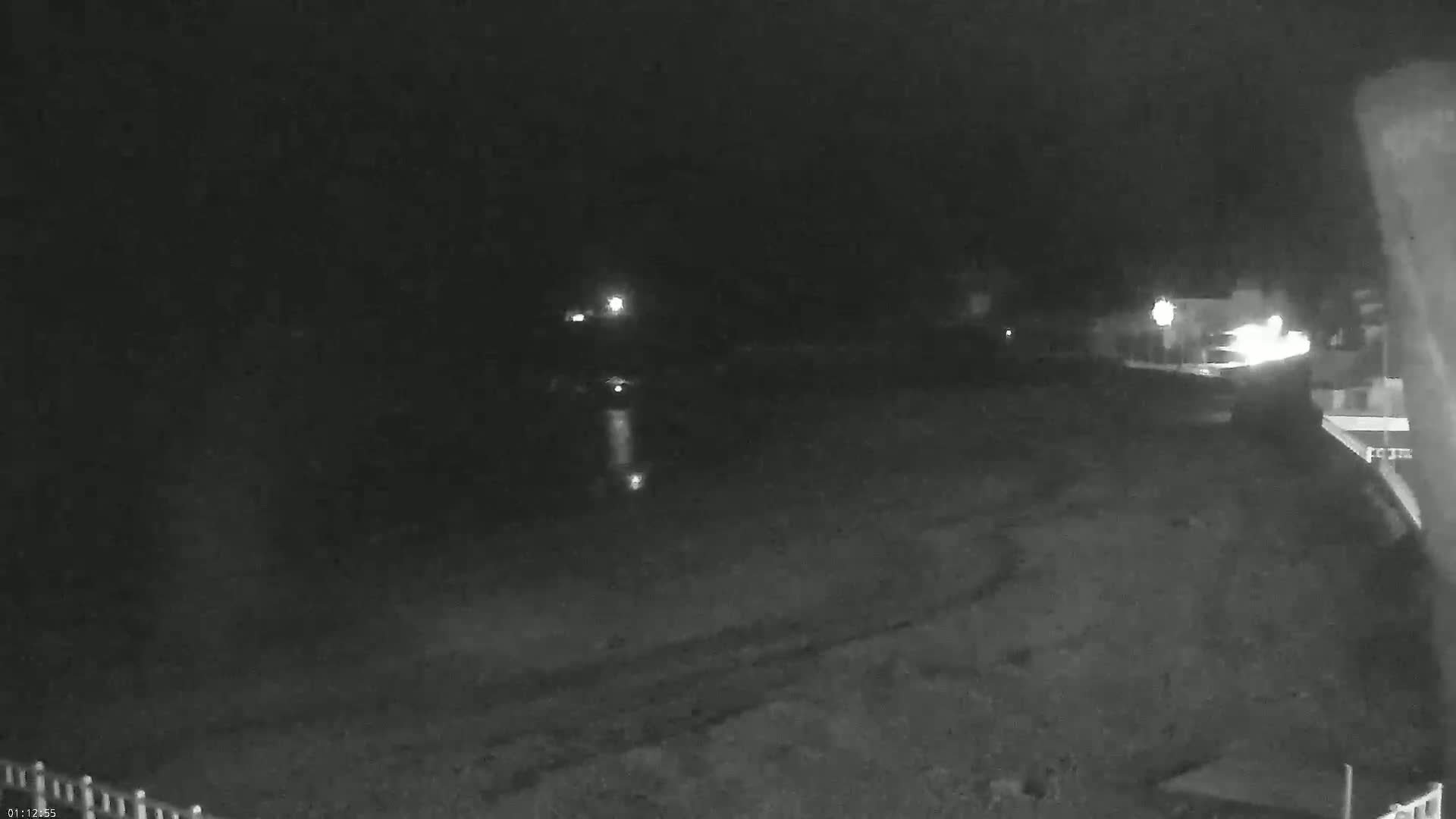 A nighttime, low-light image shows a dark, mostly empty area, possibly a beach or parking lot, with a few distant lights visible in the background.