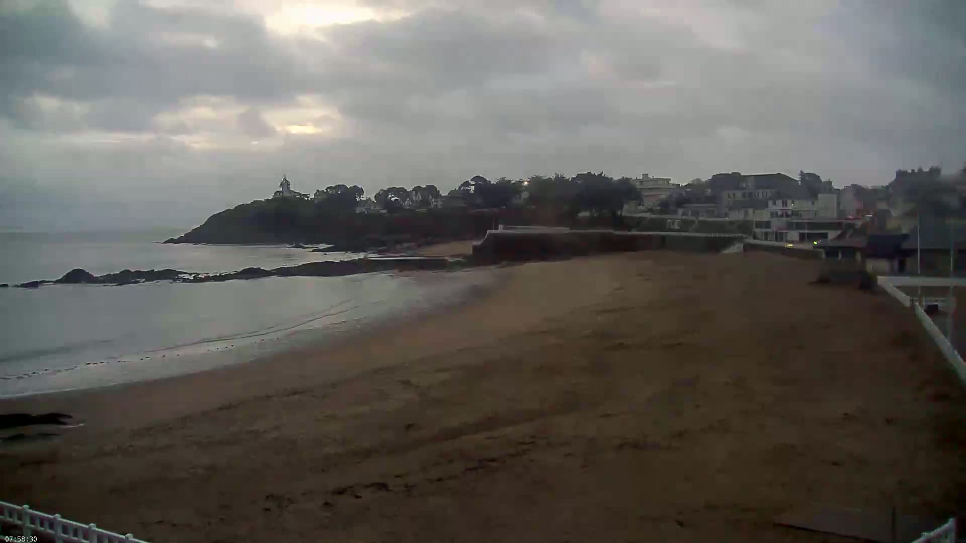 A sandy beach stretches toward a rocky coastline with buildings and trees under an overcast sky.