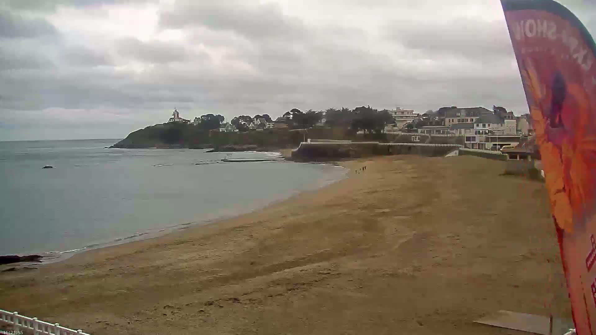 A sandy beach stretches towards a calm ocean under a cloudy sky, with a small town and a lighthouse visible on a cliff in the distance.