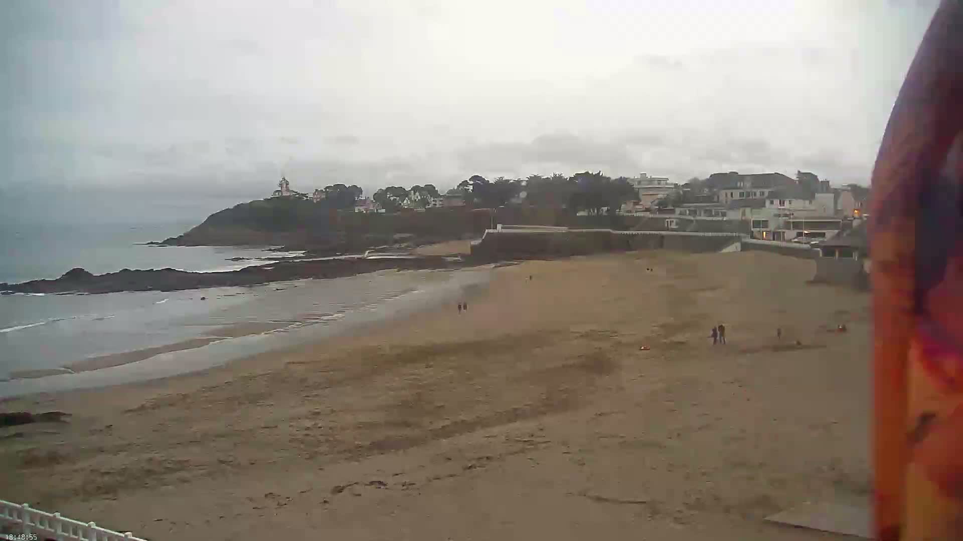 A sandy beach stretches out to a calm ocean under an overcast sky, with a small town and lighthouse visible on a nearby headland.