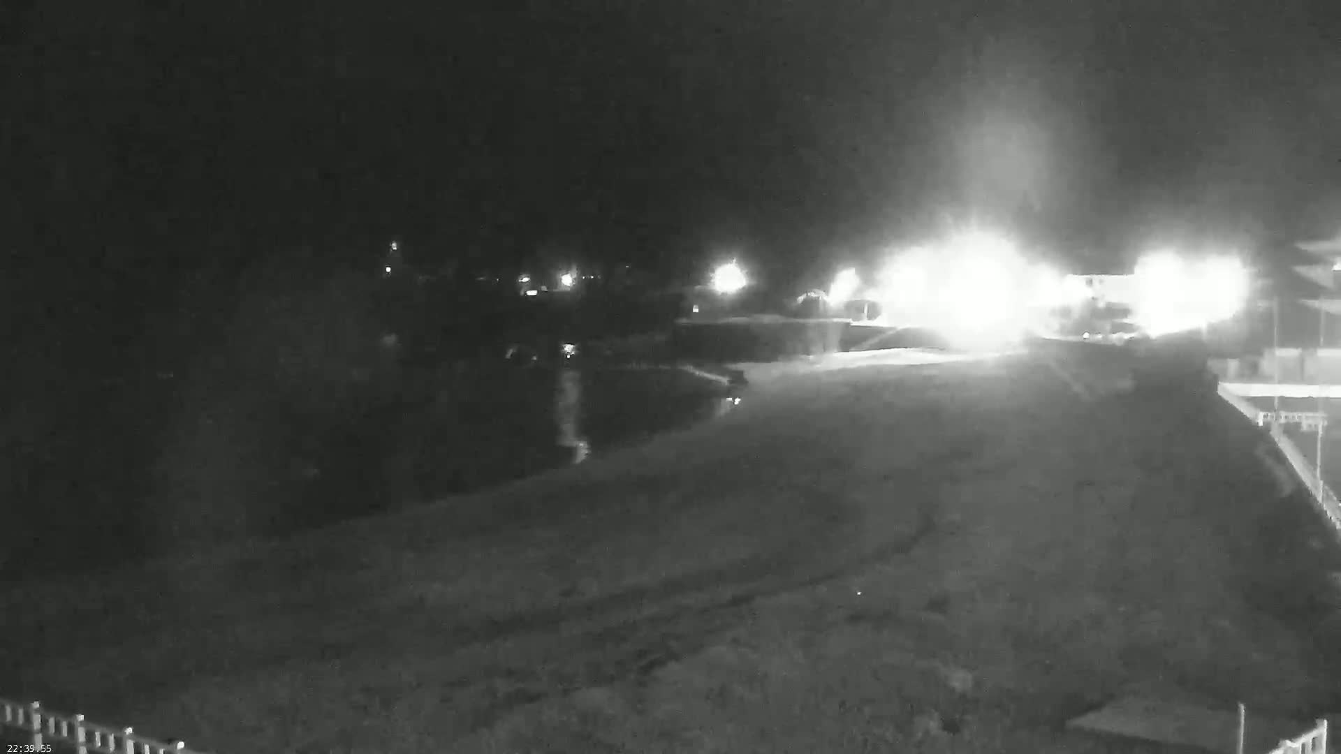 A nighttime, low-light image shows a dimly lit, empty area near a body of water with bright lights in the distance.
