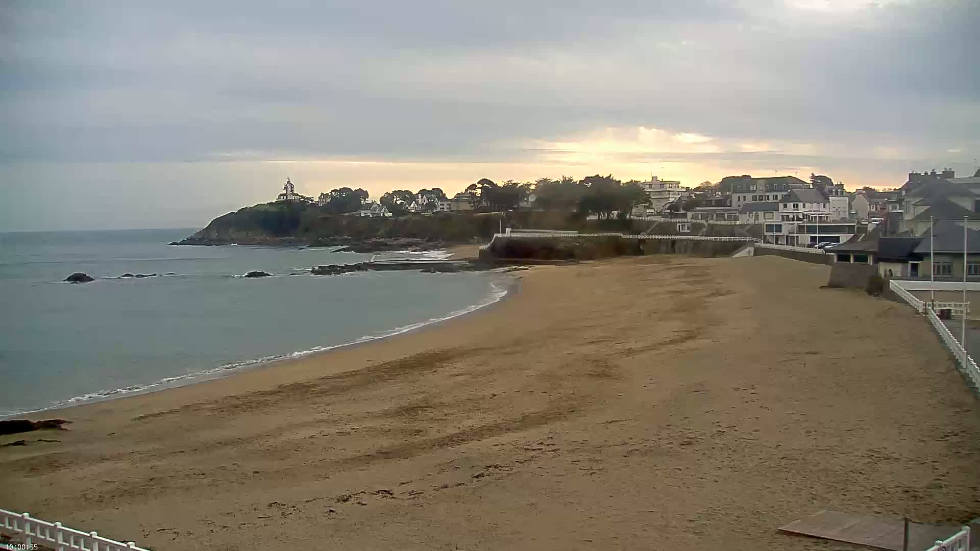 A sandy beach curves gently toward a calm ocean under an overcast sky, with buildings and a small lighthouse visible on a cliff in the distance.